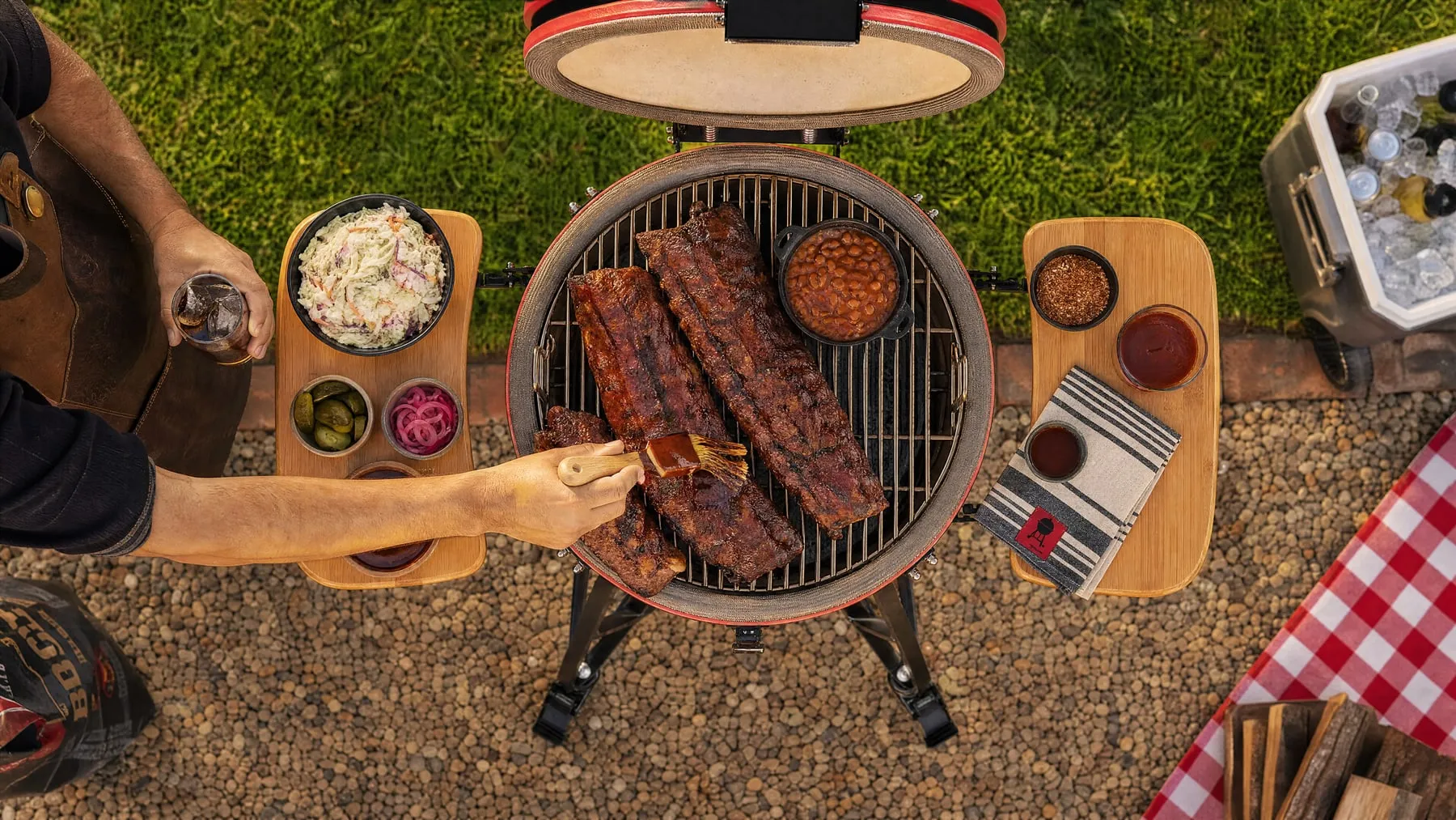 Racks of ribs on an open red kamado at a garden picnic, seen from above