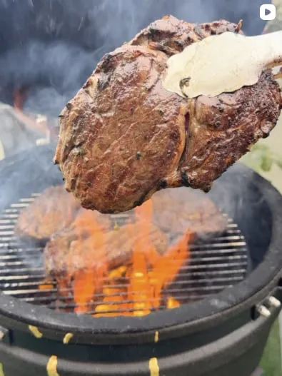 A seared steak lifted with tongs over flames on a kamado grate